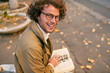 © iuricazac - Rear view of happy handsome young man reading book outdoors. College male student carrying books in campus in autumn street. Smiling smart guy wears spectacles and curly hair reading books in street