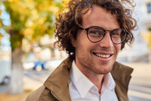 Closeup Horizontal Portrait Of Young Happy Business Man With Glasses Smiling And Posing Outdoors. Male Student In Autumn Street. Smart Guy In Casual Wears Spectacles With Curly Hair Walking On Street