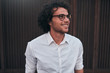 © iuricazac - Urban shot of handsome young businessman with glasses, smiling, posing outdoors. Male student posing against brown wall. Smart guy in white shirt wears spectacles with curly hair walking on street
