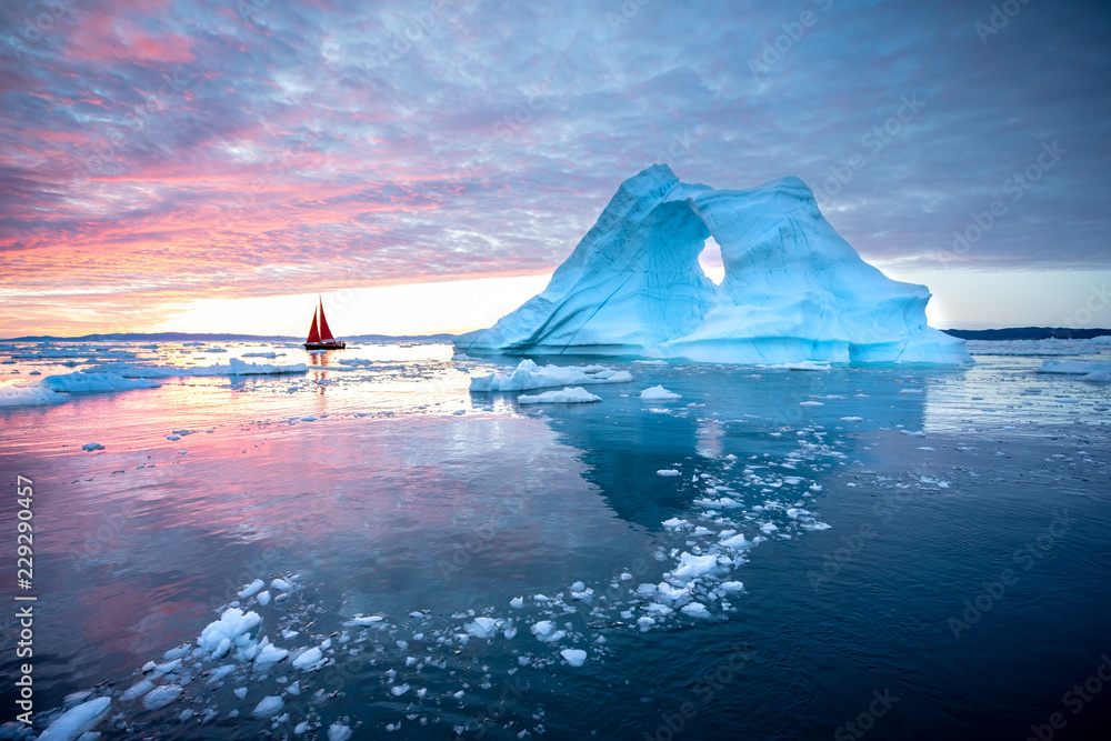 Little red sailboat cruising among floating icebergs in Disko Bay glacier during midnight sun season of polar summer. Ilulissat, Greenland. - obrazy, fototapety, plakaty 