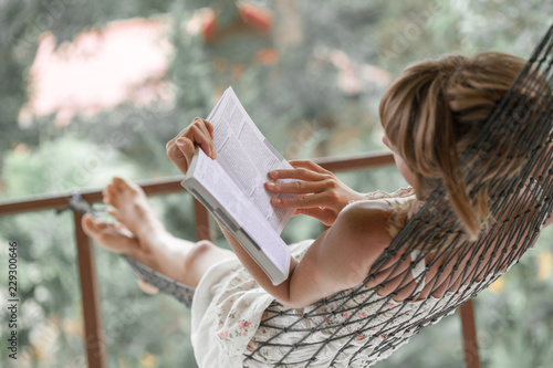 Woman in summer dress lies in the hammock in a garden and reads the book Poster Mural XXL