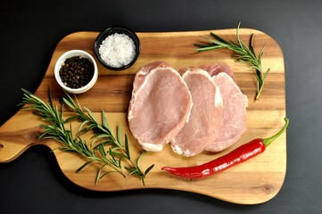 Raw fresh uncooked sliced pork fillet  dish with rosemary, pepper, salt, red chili pepper, on wooden board and black background. Top view