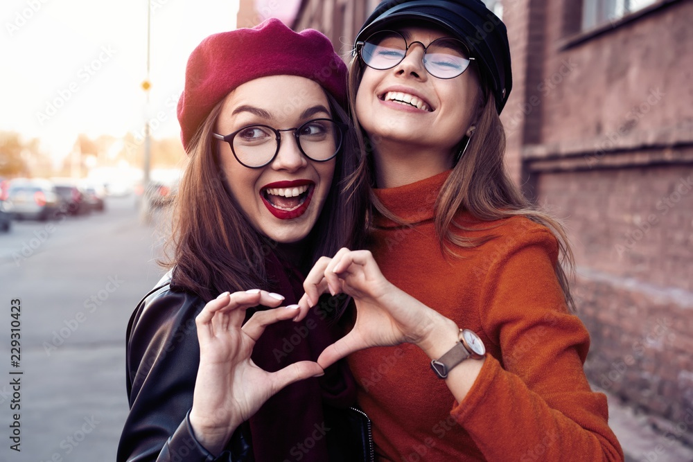 Outdoors fashion portrait young pretty best girls friends in friendly hug. Walking at the city. Posing at the street. Wearing stylish outerwear and hats. Bright make up. Positive emotions. - obrazy, fototapety, plakaty 