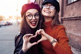 Fototapeta  - Outdoors fashion portrait young pretty best girls friends in friendly hug. Walking at the city. Posing at the street. Wearing stylish outerwear and hats. Bright make up. Positive emotions.