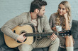 © LIGHTFIELD STUDIOS - attractive young man playing guitar for girlfriend at home while she sitting on couch with coffee
