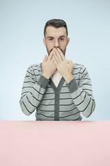 Wall Mural - Young man sitting at table covering mouth isolated on blue. I can not say anything