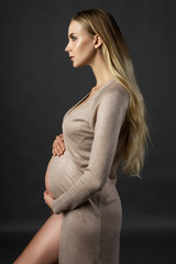 Beautiful pregnant young woman, wearing a lingerie and knitted dress, undone and open at her naked tummy softly touches her belly with her hands. Photo in profile on gray background.