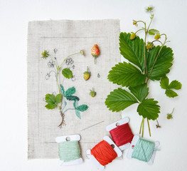 Embroidery in process with real leafs and sprig of wild strawberry and sewing threads on table.