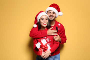 Wall Mural - Young couple in Christmas sweaters and hats with gift on color background