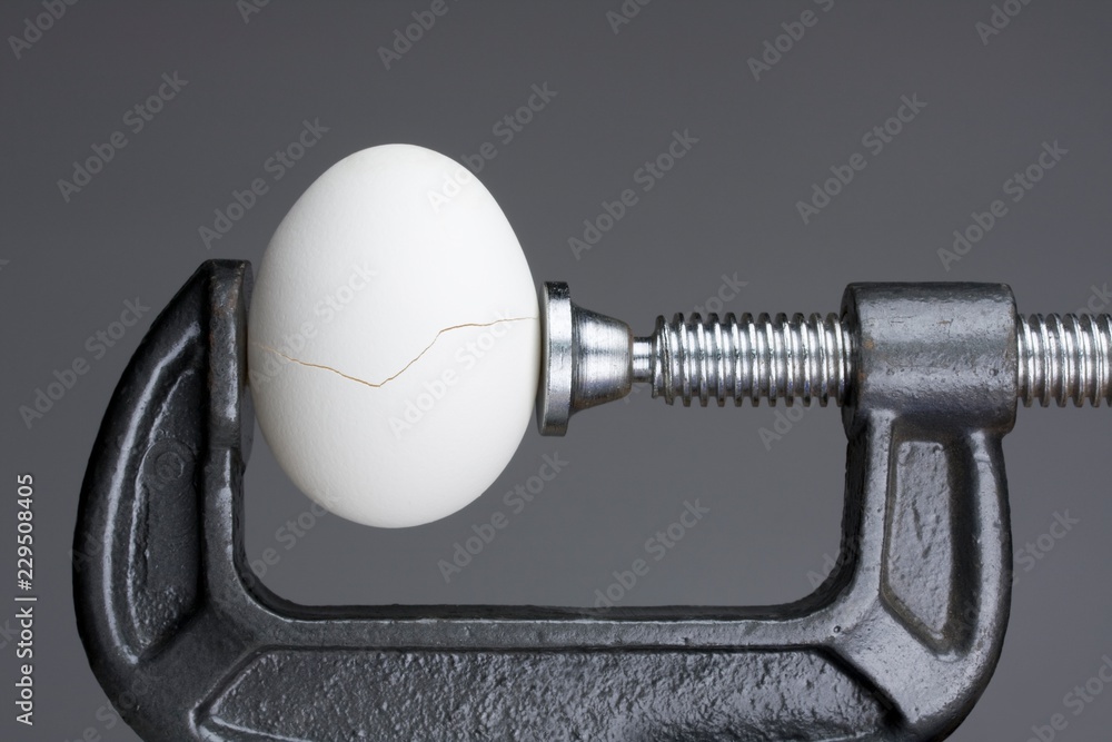 Under pressure.An egg cracking under pressure applied by squeezing clamps form the sides. - obrazy, fototapety, plakaty 