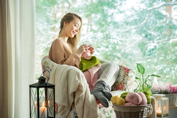 Young woman sitting home in a chair by the window wearing knitted warm sweater, knitting with needles. Cozy room decorated with lanterns and candles. Scenic winter view of pine trees in snow in window