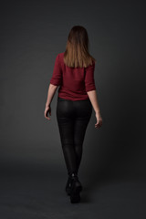 Wall Mural - full length portrait of brunette girl wearing  red shirt and leather pants. standing pose, facing away from the camera, on grey studio background.