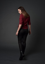Full Length Portrait Of Brunette Girl Wearing  Red Shirt And Leather Pants. Standing Pose, Facing Away From The Camera, On Grey Studio Background.