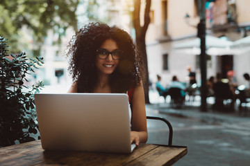 Cheerful young woman freelancer is working on her laptop in an outdoor cafe; smiling caucasian female with curly hair using the netbook while sitting at the wooden table of a street bar, Barcelona