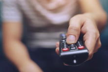 Close Up Television Remote Control In Casual Man Hands Pointing To Tv Set And Turning It On Or Off. Select Channel Watching Tv On His Sofa At Home In The Living Room Relax.