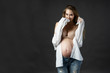 Beautiful smiling pregnant young blond woman, wearing ripped jeans, bra and unbuttoned white shirt, stays and touches her face. Naked belly. Gray background. Lifestyle, pregnancy, health