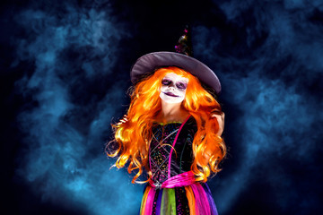  Beautiful girl in a witch costume on a dark background in smoke. Portrait of little girl in carnival costume of sorceress, background on halloween.
