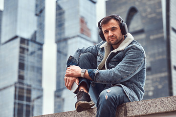 Stylish hipster man trendy dressed listening to music sitting in front of skyscrapers in Moskow city at cloudy morning.