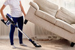 Portrait of young woman in white shirt and jeans cleaning carpet under sofa with vacuum cleaner in living room, copy space. Housework, cleanig and chores concept