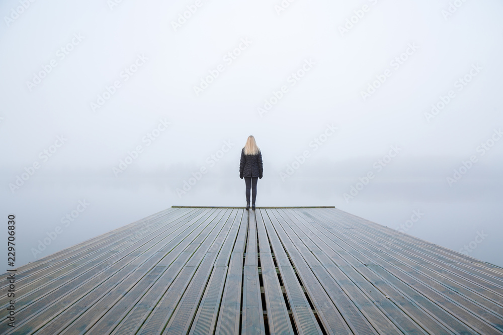 Young woman standing alone on edge of footbridge and staring at lake. Mist over water. Foggy air. Early chilly morning in autumn. Beautiful freedom moment and peaceful atmosphere in nature. Back view. - obrazy, fototapety, plakaty 