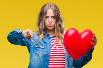 Wall Mural - Beautiful young blonde woman holding heart valentine over isolated background with angry face, negative sign showing dislike with thumbs down, rejection concept