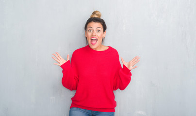 Young adult woman over grey grunge wall wearing winter outfit celebrating crazy and amazed for success with arms raised and open eyes screaming excited. Winner concept