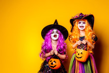 Two Beautiful Girls In A Witch Costumes And Hats On A Yellow Background Scaring And Making Faces. Portrait Of Little Girls In Carnival Costumes Of Sorceress, Background On Halloween.