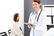 © rogerphoto - Happy doctor woman at work. Portrait of female physician using tablet computer while standing near reception desk at clinic or emergency hospital. Patient woman sitting at the background. Medicine