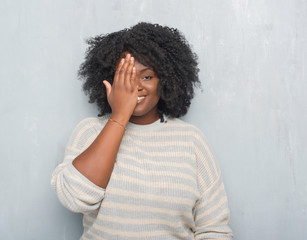 Sticker - Young african american plus size woman over grey grunge wall wearing a sweater covering one eye with hand with confident smile on face and surprise emotion.