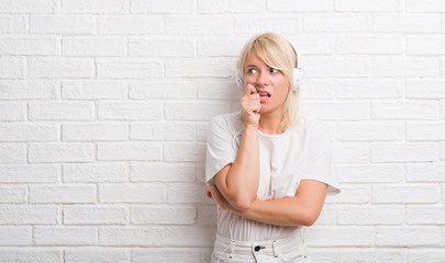 Poster - Adult caucasian woman over white brick wall wearing headphones looking stressed and nervous with hands on mouth biting nails. Anxiety problem.