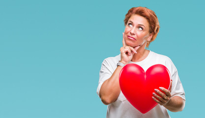 Wall Mural - Senior caucasian woman holding red heart in love over isolated background serious face thinking about question, very confused idea