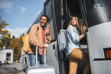 smiling multiracial male tourist with wheeled bag waving by hand while his girlfriend walking into travel bus at city street