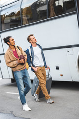 Wall Mural - multiracial young man with rugby ball walking with male friend carrying bag on wheels near bus at city street