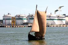An Ancient Boat With Sails On The Background Of A Port Oil Terminal