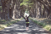 Blonde Female Raises Arms Up At The Cypress Tree Tunnel On Point Reyes National Seashore In Marin County In Northern California