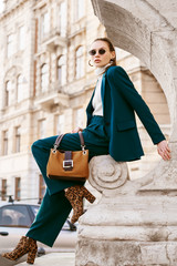 Outdoor full body fashion portrait of young woman wearing sunglasses, white turtleneck, blue suit, blazer, trousers, leopard print ankle boots, holding suede bag, posing in street of european city