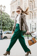 Outdoor full body fashion portrait of  fashionable woman wearing sunglasses, white turtleneck, leopard print blazer, boots, green trousers, holding brown suede bag, walking in street of european city