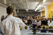 © Valmedia - Man with white shirt is giving a conference in front of 200 people in an industrial environment with yellow and white walls - Blurred audience mainly composed of young adults