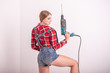 © Михаил Решетников - A young sexy woman stands with her back near the white wall and holds a drill. The girl in shorts and a plaid shirt makes repairs using a perforator. woman makes repairs with a tool.