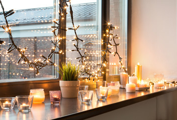 hygge, decoration and christmas concept - candles burning in lanterns and festive garland on window 