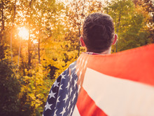 Attractive, Stylish Man Holding A Waving US Flag On The Background Of The Rays Of The Setting Sun And Yellow Trees. View From The Back. Preparing For The Holidays