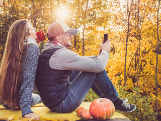 Attractive man and stylish woman holding a mobile phone and sitting on the terrace against the background of yellow trees and the setting sun. Happy relationship concept