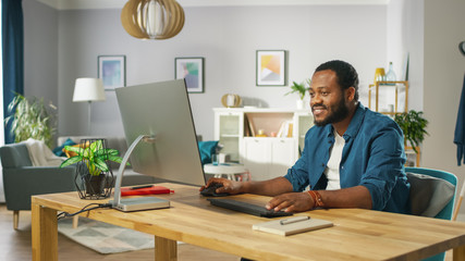 handsome black man works on a personal computer while sitting at the desk of his cozy living room.