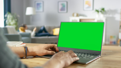 Wall Mural - Over the Shoulder Shot of Mans Hands Typing on a Laptop with Green Mock-up Screen. In the Background Cozy Living Room with Woman Relaxing on a Sofa.