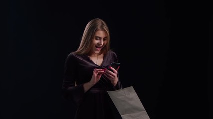 Wall Mural - Woman using phone purchase in black dress, holding black shopping bag isolated on dark background in black friday holiday or christmas