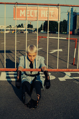 fashionable portrait of woman sitting on asphalt with white marking road near metal fence, arms between bars, black lace gloves, whit short hair, black tie, looking down, parking lot