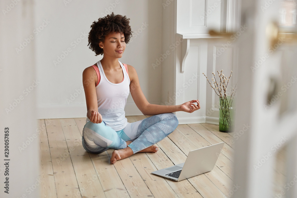 Relaxed dark skinned woman with sporty body, sits in zen pose, keeps legs crossed, watches yoga lessons on laptop computer using internet, meditates on floor in empty room. Wellness concept. - obrazy, fototapety, plakaty 