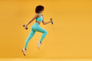 Wall Mural - Strong athletic, woman sprinter or runner, running on yellow background with dumbbells wearing sportswear. Fitness and sport motivation.