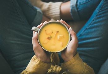 Wall Mural - Flat-lay of female in yellow shirt and jeans sitting and keeping mug of Fall warming yellow pumpkin cream soup with croutons, top view. Autumn vegetarian, vegan, healthy comfort food eating concept