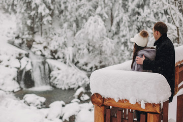 Stylish couple hugging and looking at snowy woods from wooden porch in mountains. Happy family relaxing on background of winter mountains. Holiday getaway together. Space for text